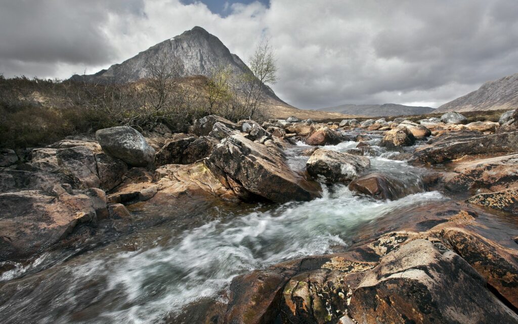Rushing river flows over rocks below towering mountain under dramatic gray cloudy sky wallpaper