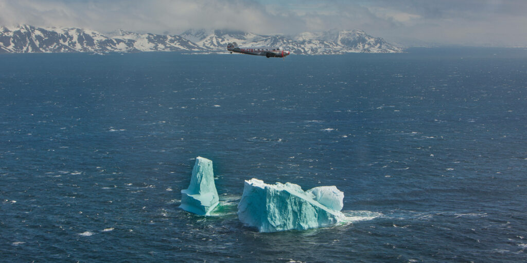 Lockheed Electra plane flying over icy blue Greenland waters with icebergs and snowy mountains Wallpaper