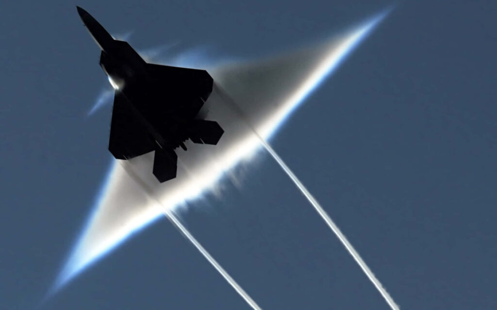Lockheed Martin F-22 Raptor jet breaking sound barrier with vapor cones in clear blue sky Wallpaper