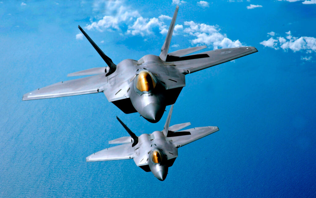 Two Lockheed Martin F-22 Raptor fighter jets flying over blue ocean with gold cockpit canopies Wallpaper