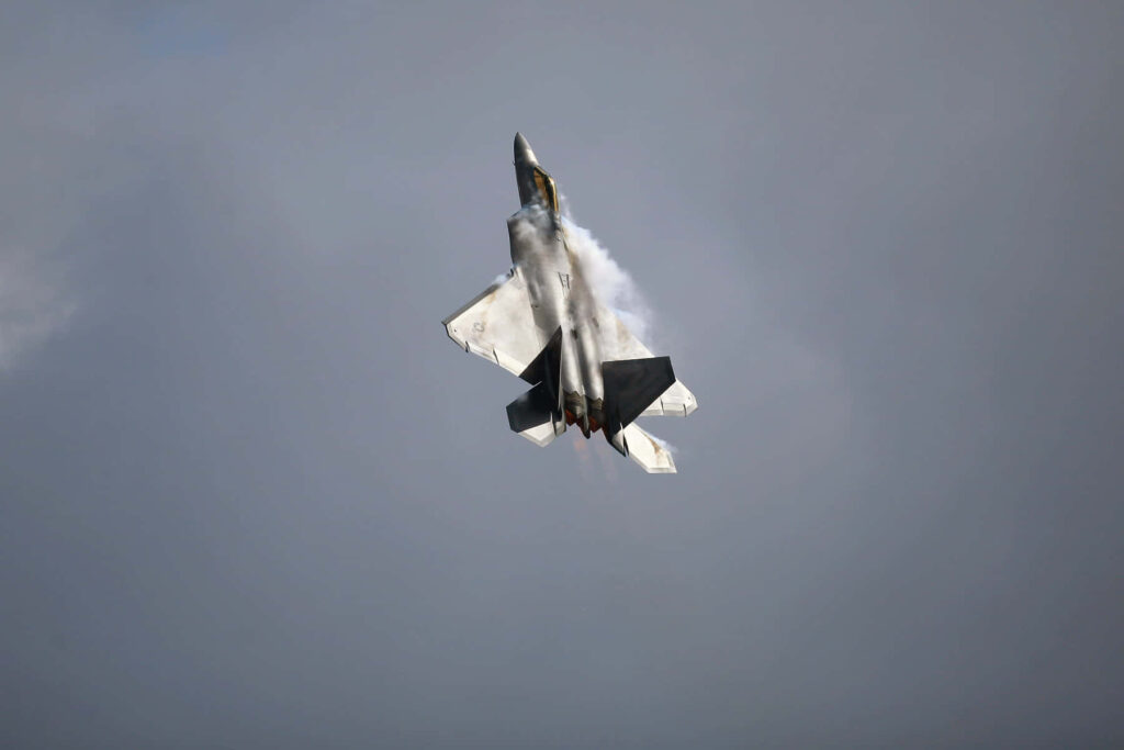 Lockheed Martin F-22 Raptor stealth fighter in vertical climb with vapor trails against cloudy sky Wallpaper