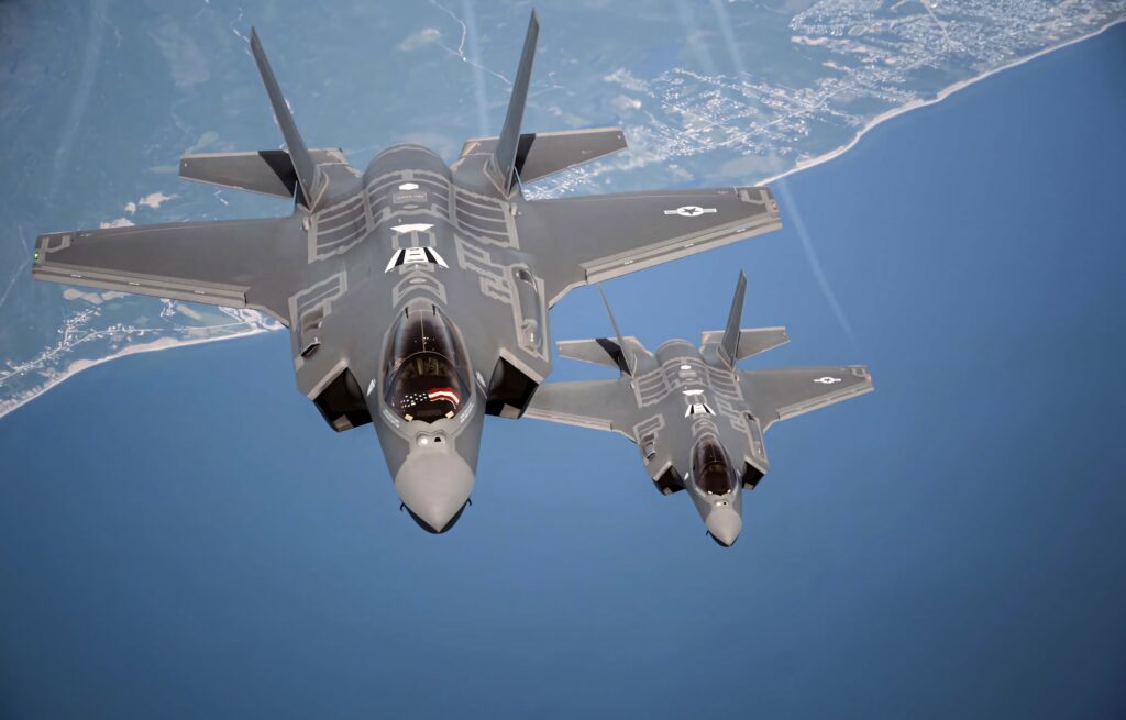 Two Lockheed Martin F-35 fighter jets flying over coastline with blue ocean and landscape background Wallpaper