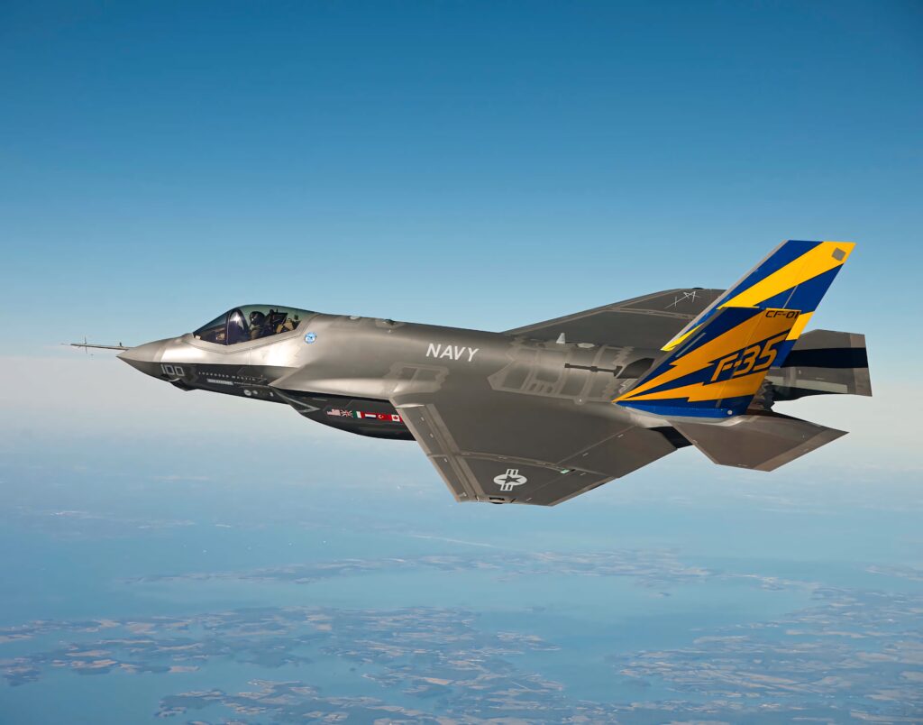 Lockheed Martin F-35 jet soaring over blue terrain with metallic gray body and yellow-blue tail fin Wallpaper