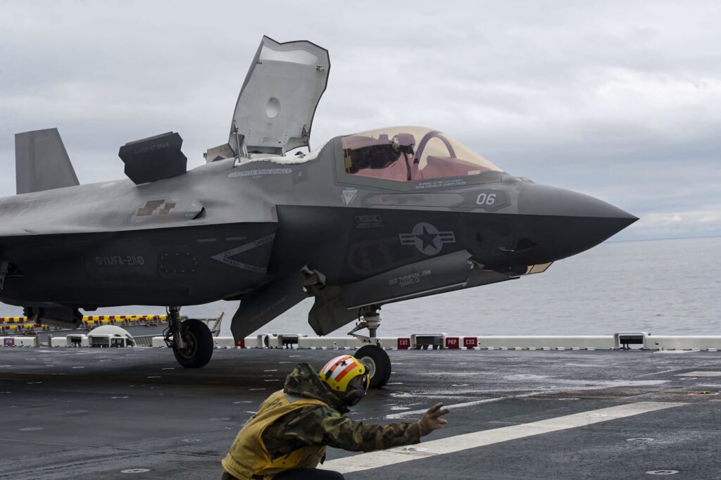 Lockheed Martin F-35 stealth fighter jet on aircraft carrier deck with deck crew signaling under gray skies Wallpaper
