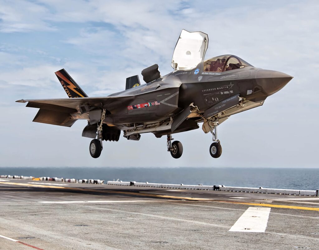 Lockheed Martin F-35 stealth jet hovering above aircraft carrier deck with ocean and sky backdrop Wallpaper