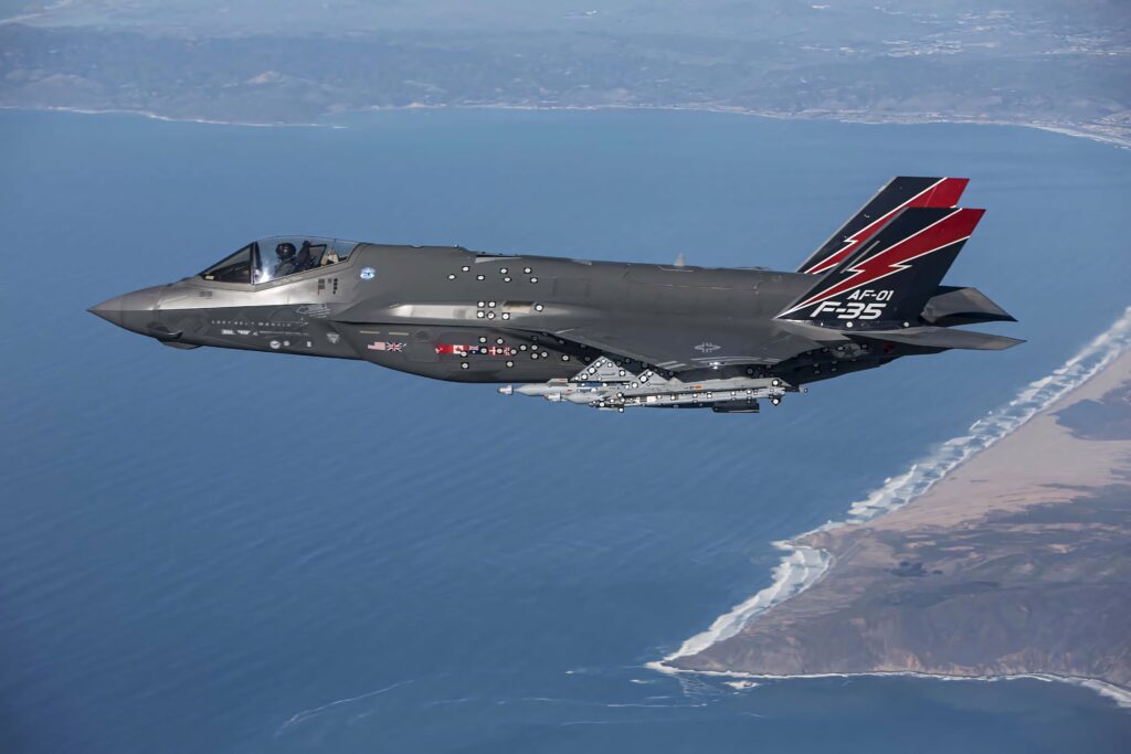 Lockheed Martin F-35 stealth jet flying over coastal landscape with red black tail markings wallpaper