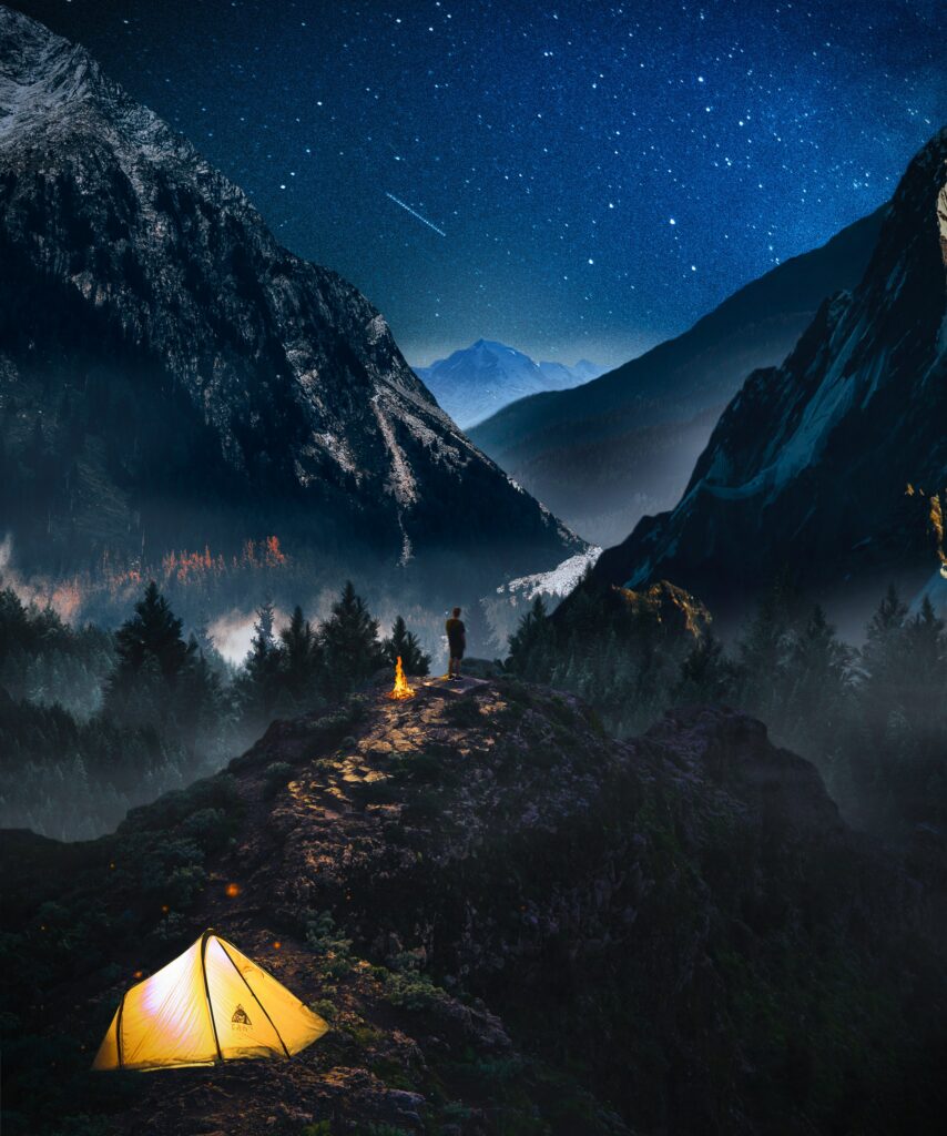 Lone camper on rocky ridge under starry sky with glowing tent campfire in mountain forest wilderness Wallpaper