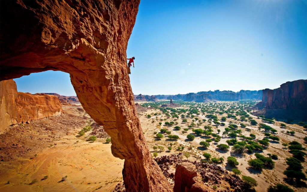 Lone climber ascending massive rock formation under bright blue sky with sunlit desert background Wallpaper