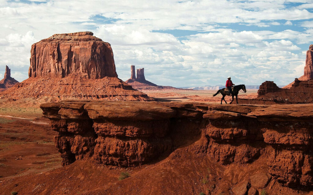 Lone cowboy rides horse along red sandstone cliff with towering mesas under cloudy sky Wallpaper