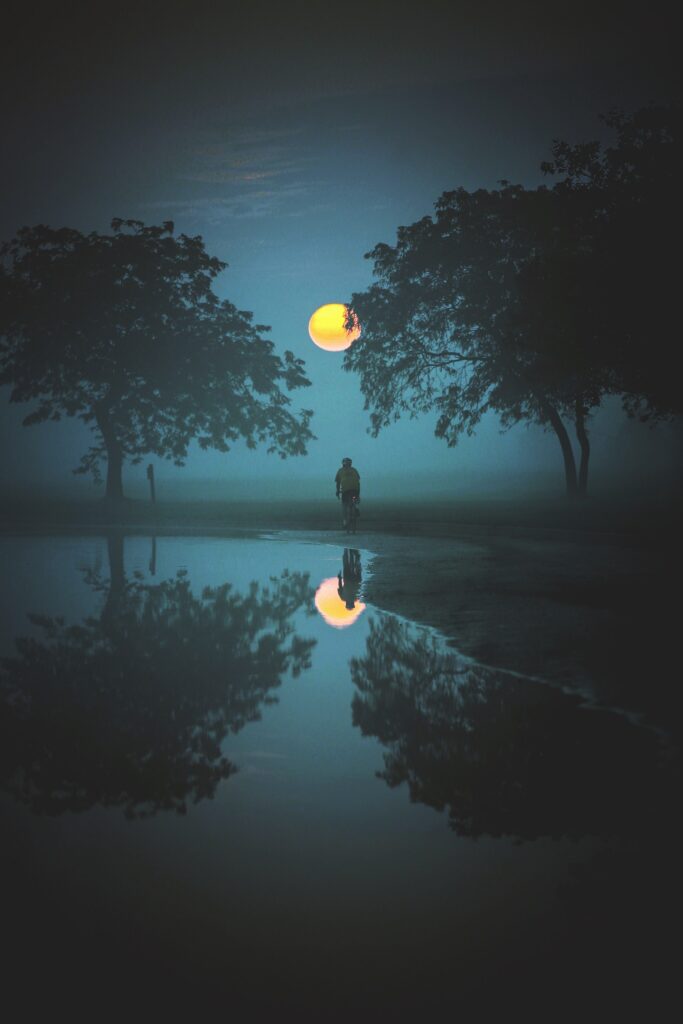 Lone cyclist rides through fog under full moon framed by trees reflected in calm water wallpaper