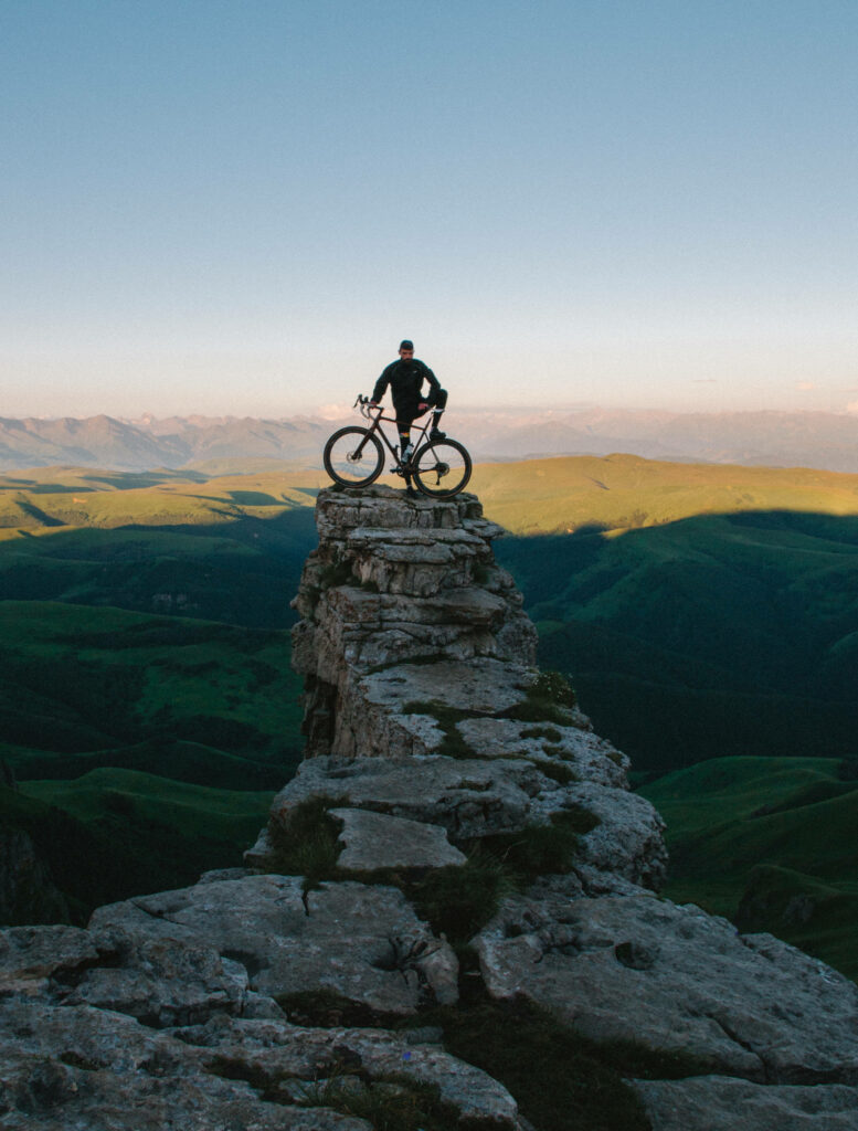 Lone cyclist on rock cliff overlooking green hills under clear blue sky adventure and freedom Wallpaper