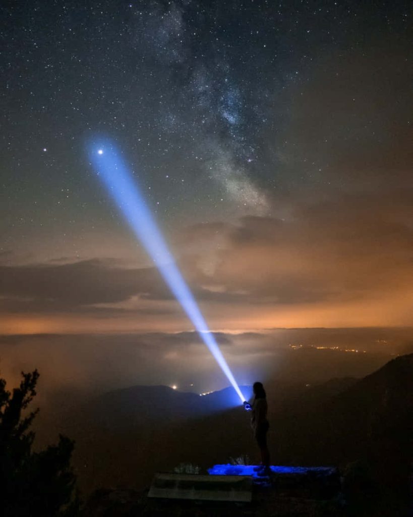 Silhouette with blue flashlight on mountain under Milky Way stars city lights night wallpaper