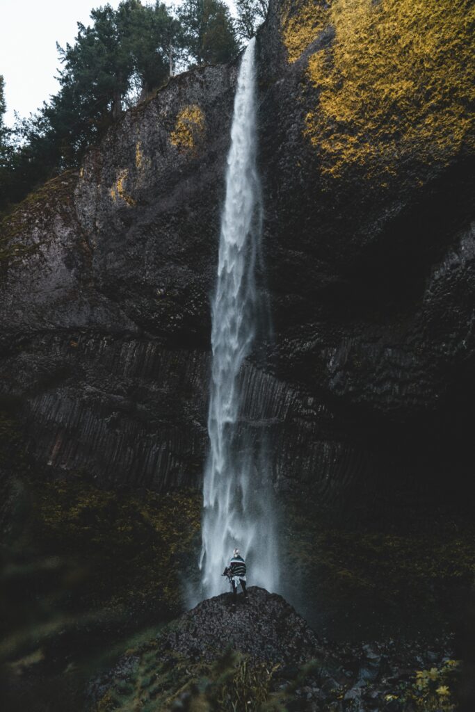 Lone person on rocky cliff under narrow waterfall among mossy yellow cliffs dramatic nature wallpaper