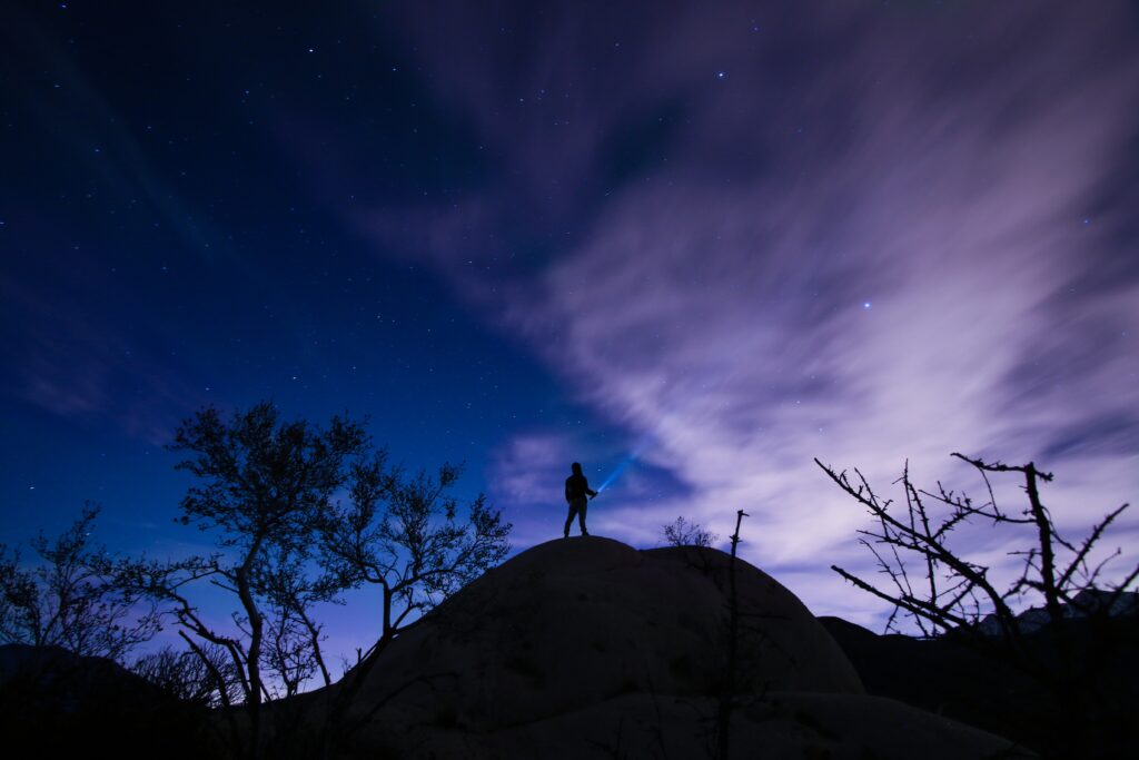 Lone figure on hill beneath starry sky with silhouetted trees and dramatic night clouds wallpaper