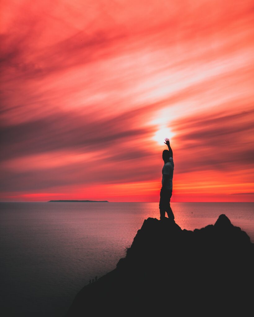 Silhouetted man on rugged hill by sea at vivid red sunset with calm water and vast horizon wallpaper