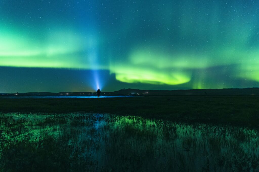 Man admiring vibrant green and blue northern lights reflected on calm water under night sky Wallpaper
