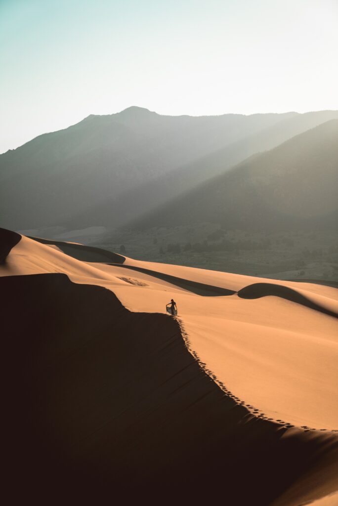 Man walking on golden sand dunes leaving footprints in sunlit desert with distant mountains Wallpaper