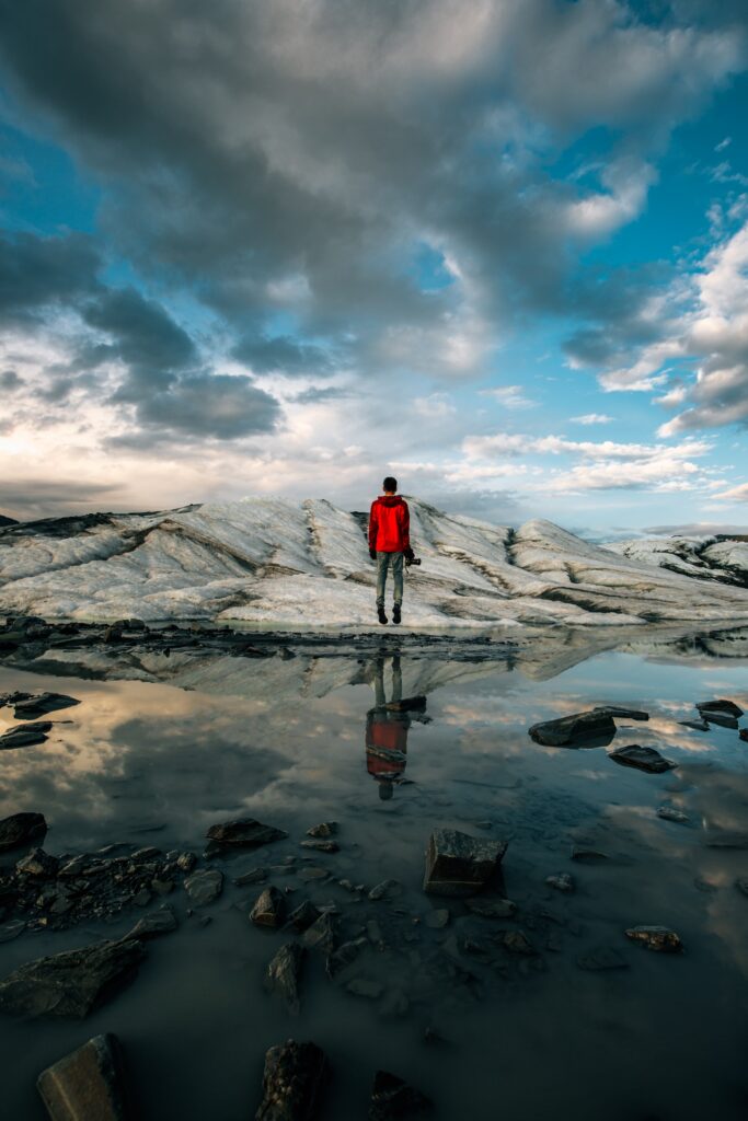 Man in red jacket facing vast glacier reflected in calm water under dramatic blue sky outdoors Wallpaper