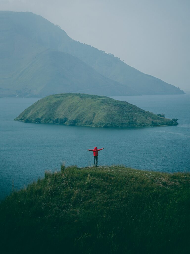 Man in red jacket on grassy cliff overlooking misty blue sea island and foggy mountains wallpaper