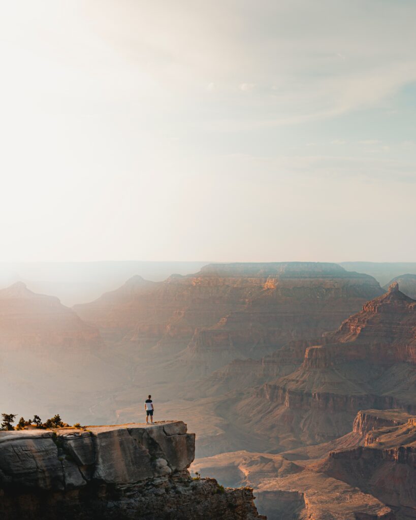 Man on cliff edge overlooking sunlit mountains and layered rock formations at sunrise Wallpaper