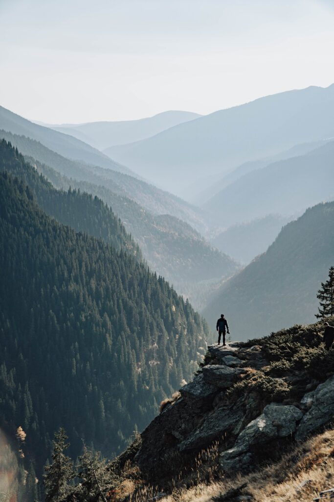 Man standing on rocky outcrop overlooking misty mountain ranges and dense pine forests wallpaper