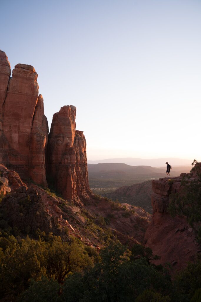 Man on rugged canyon ledge with red rocks and green trees under clear sky landscape wallpaper