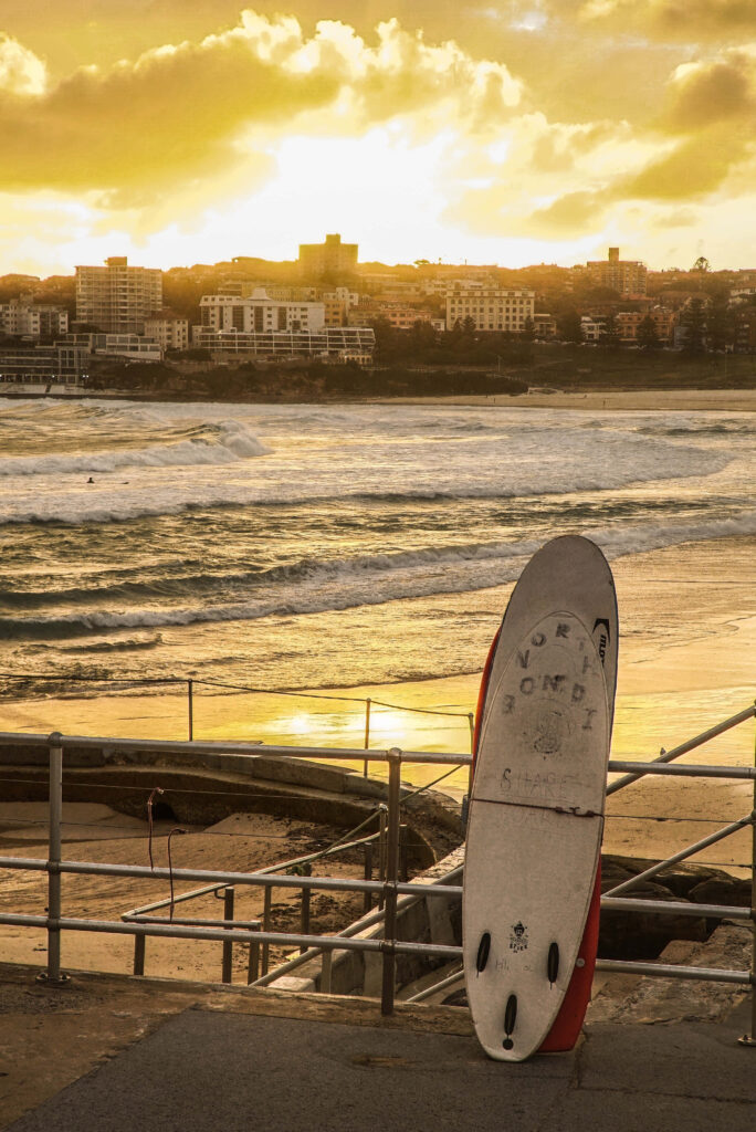 Lone Surfboard at Sunset: Serene Coastal Scene with Cityscape Wallpaper