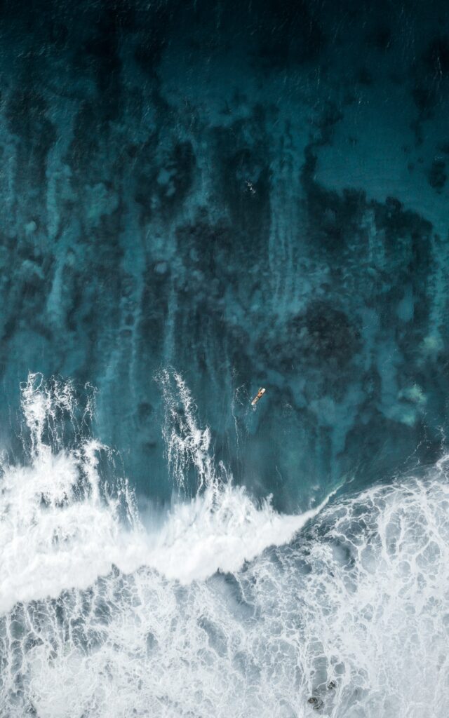 Aerial view of lone surfer paddling through foamy waves in deep blue ocean wallpaper background