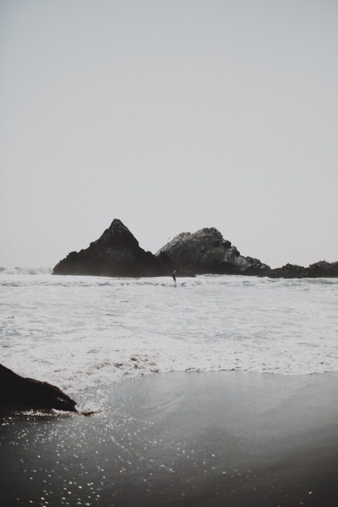 Surfer riding gentle waves near rocky coast under pale sky capturing serene shoreline vibe Wallpaper