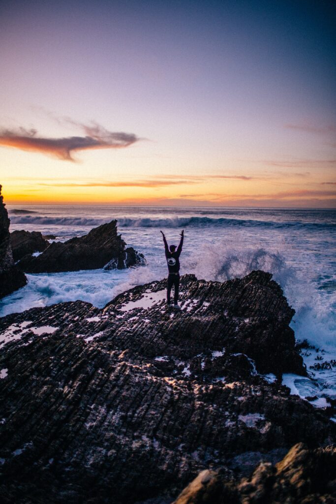 Surfer stands on rugged rocks facing crashing waves under vibrant sunset sky with blue purple hues Wallpaper
