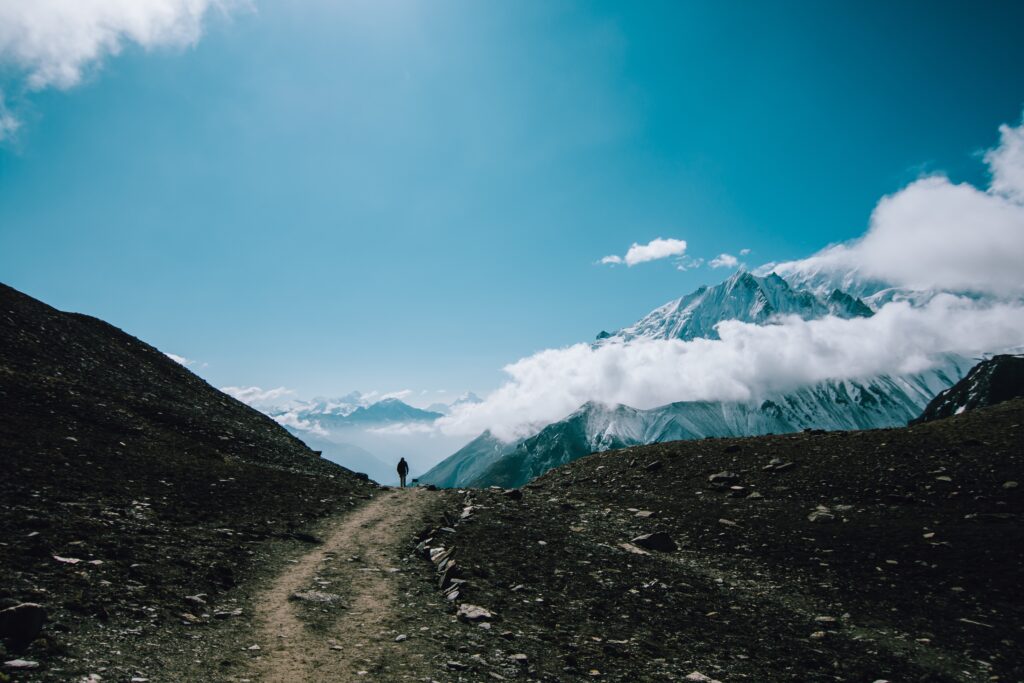 Lone tourist hiking rugged mountain trail with dramatic peaks and rolling clouds under blue sky Wallpaper