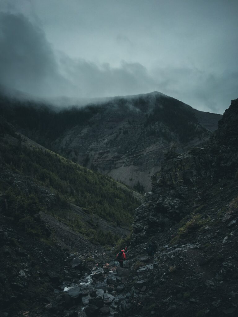 Lone tourist in red jacket hiking rugged foggy mountain landscape with river and dark stones Wallpaper