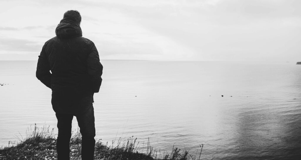 Solitude at the Shore: A Black and White Silhouette of a Man Standing Alone near the Body of Water as a Wallpaper Background Photo