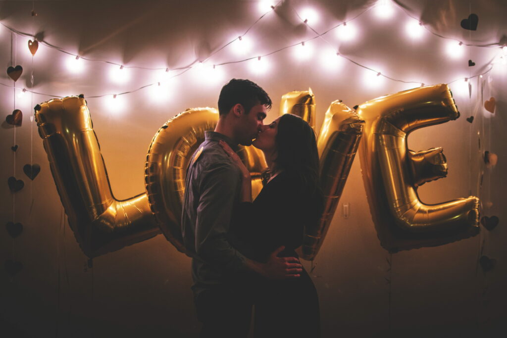 Romantic couple kissing under warm string lights with giant gold LOVE balloons and hearts background Wallpaper
