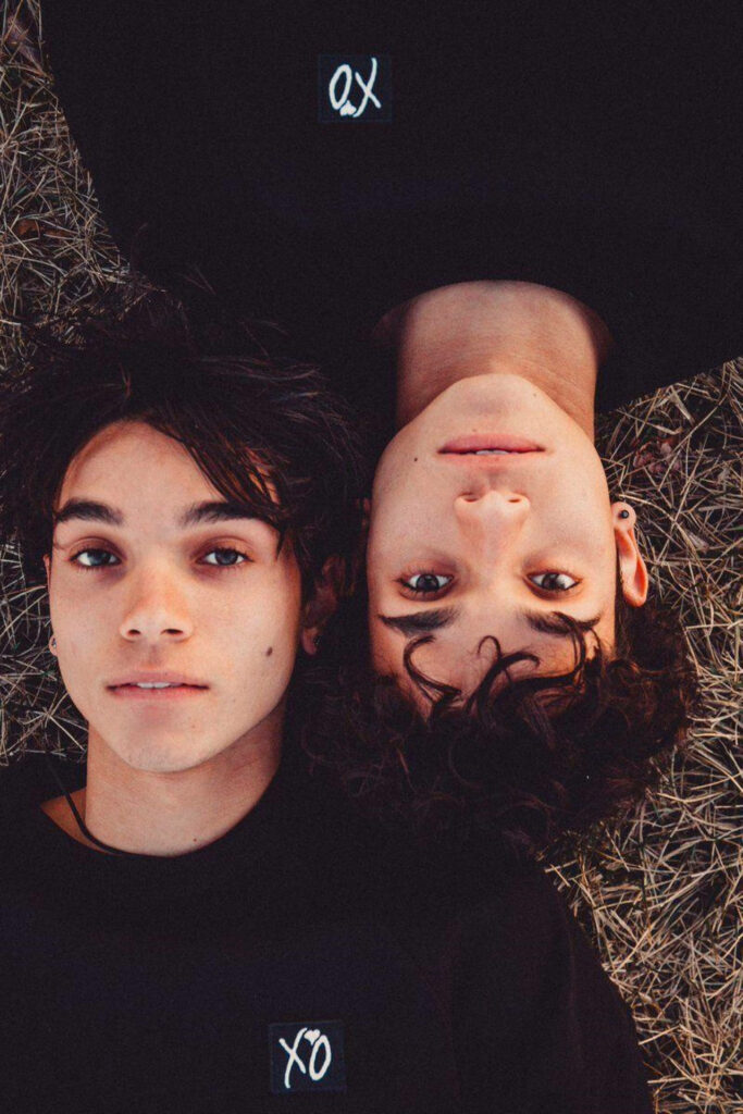 Lucas and Marcus Dobre Brothers lying head to head on earthy toned wallpaper background in black outfits