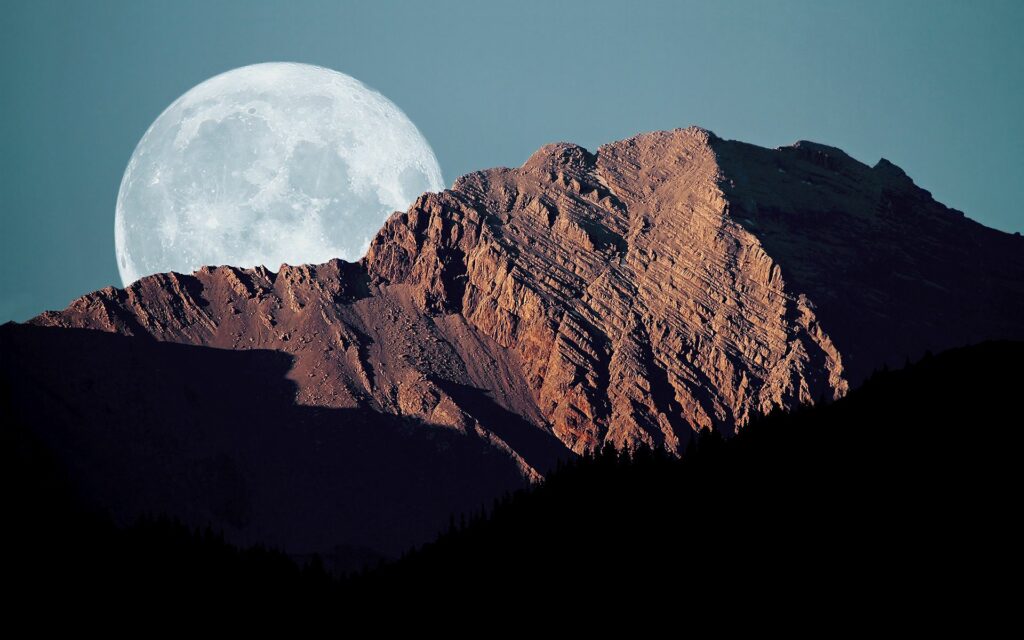 Full moon rising behind jagged mountains casting shadows on rugged rocks under deep blue sky Wallpaper