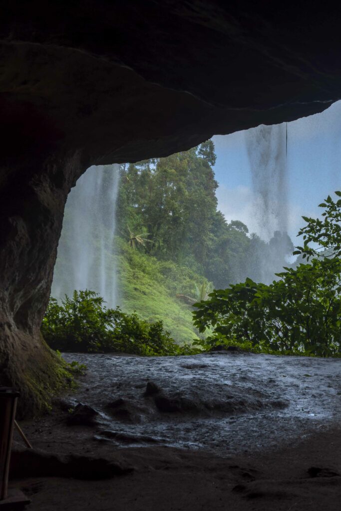 View from cave opening to vibrant forest with waterfalls lush green trees and misty spray Wallpaper