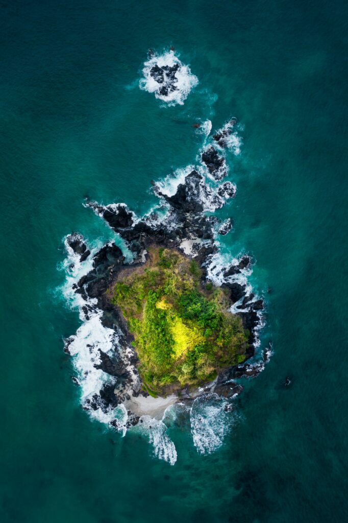 Aerial view of lush green island with rugged black rocks and deep blue ocean waves crashing coast Wallpaper