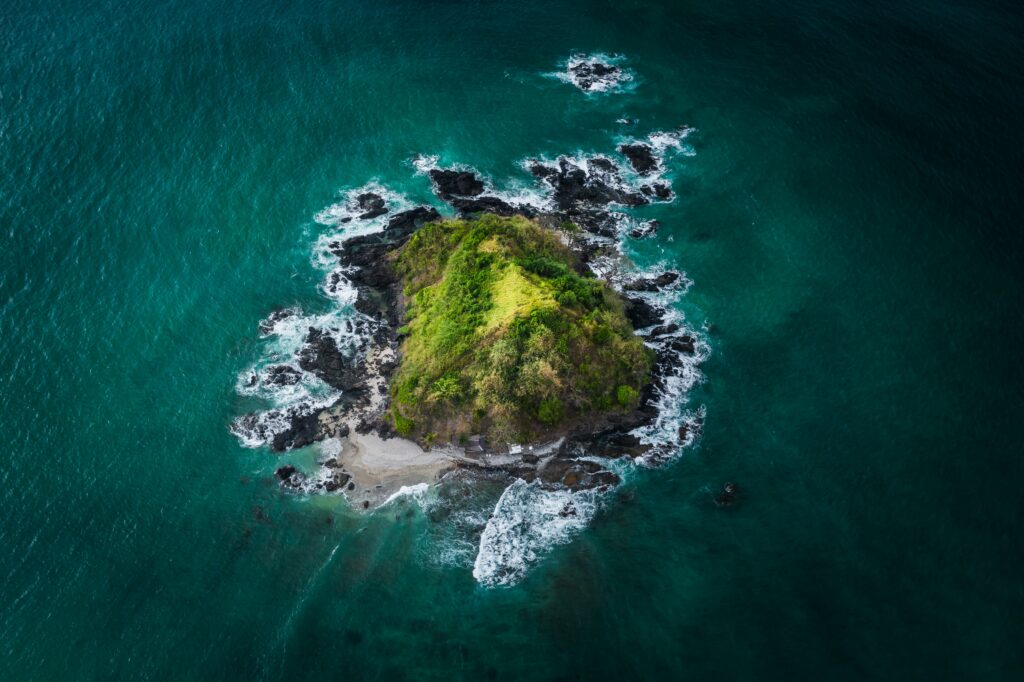 Aerial view of lush green island surrounded by black volcanic rocks and turquoise ocean waves Wallpaper