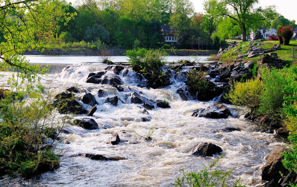 Lively river cascading over dark rocks with lush greenery houses and tall trees in soft daylight Wallpaper