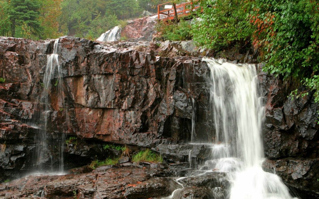Cascading waterfalls over rugged rocks with lush green foliage and a wooden bridge view Wallpaper