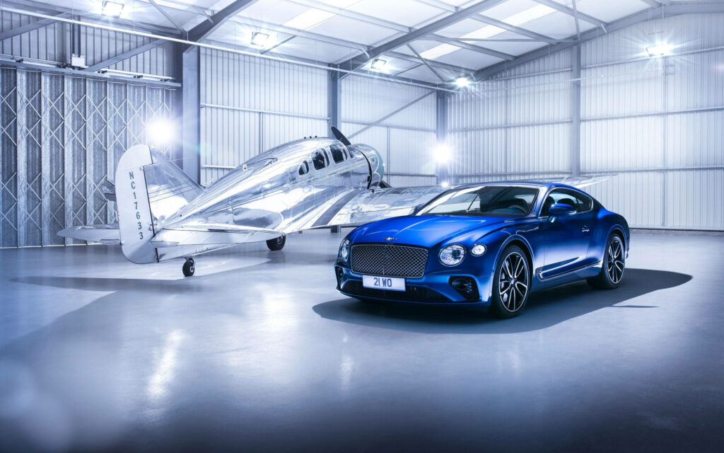 Luxury on Land and Air: Blue Bentley Continental GT Beside a Gleaming Silver Airplane in a Radiant Room Wallpaper