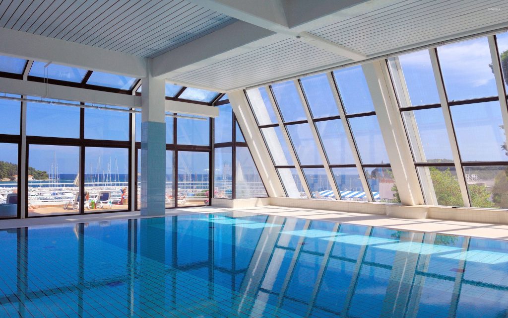 Indoor pool with glass walls overlooking marina sailboats under bright natural light Wallpaper