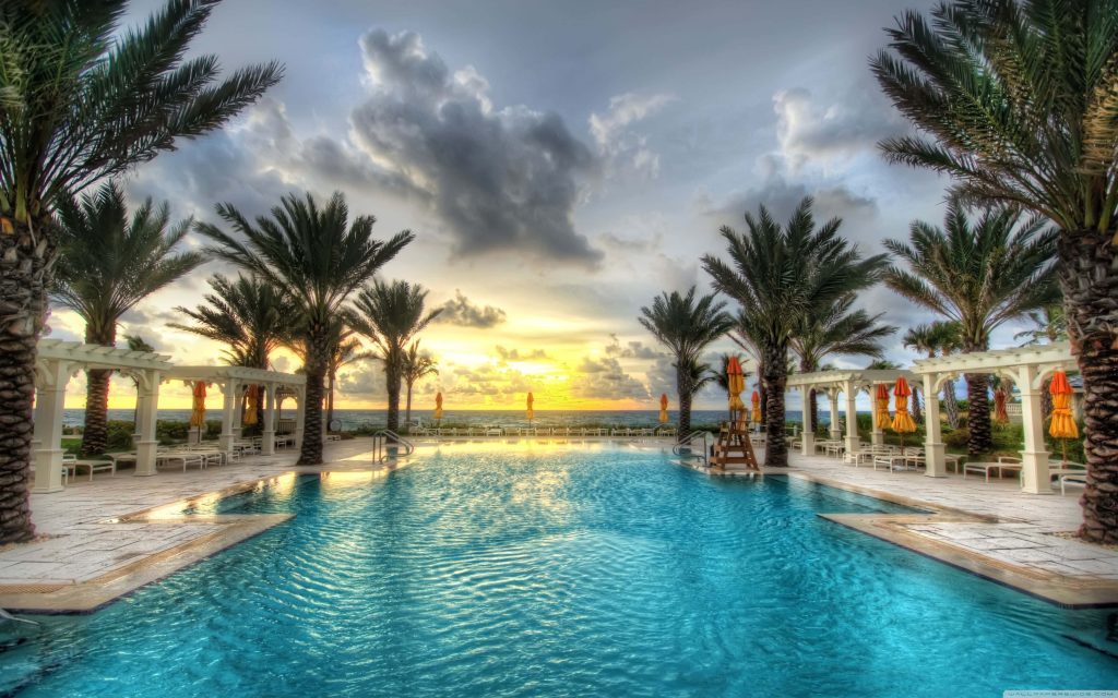 Luxurious oceanfront pool with palm trees and white pergolas reflecting vibrant sunset sky wallpaper