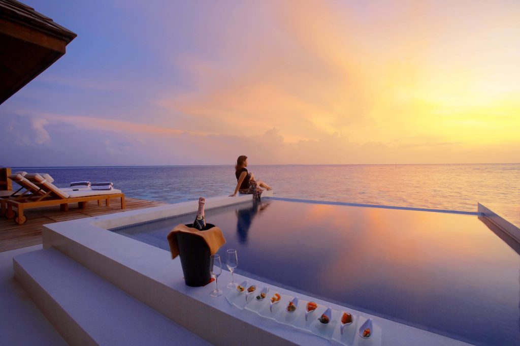 Woman relaxing by infinity pool at overwater villa with sunset ocean and Moët champagne luxury getaway Wallpaper
