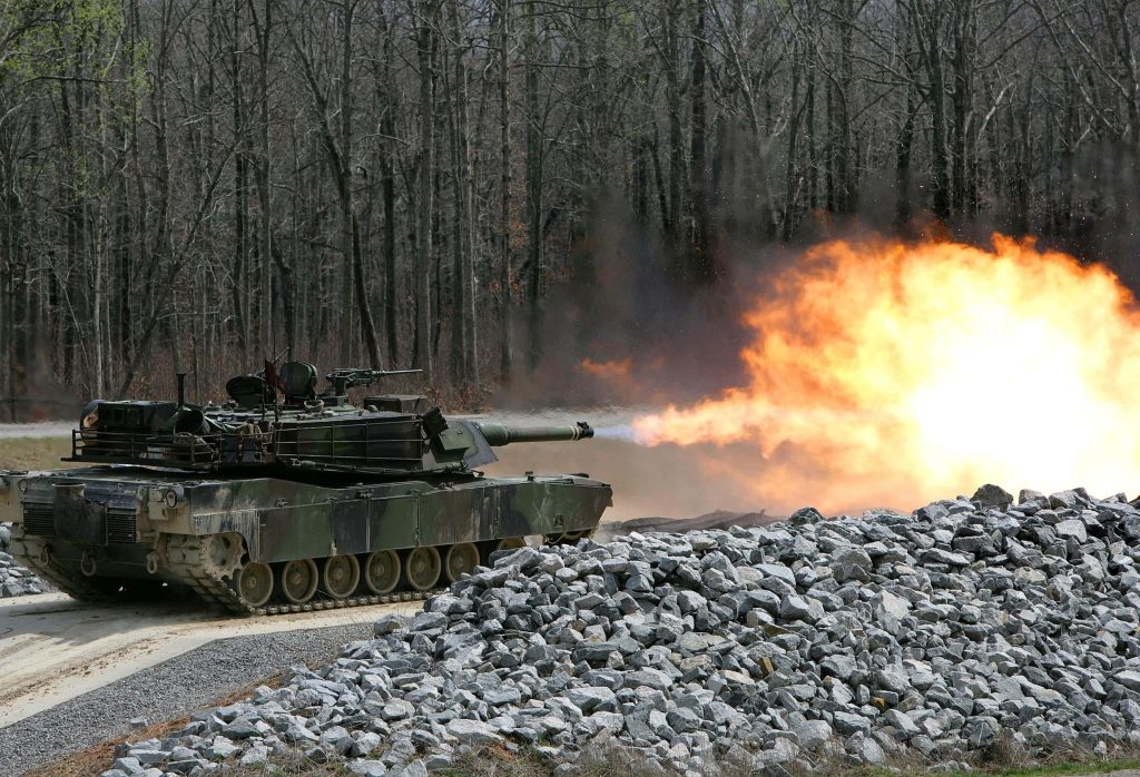 M1 Abrams tank firing cannon with bright muzzle flash in rugged forested battlefield background Wallpaper