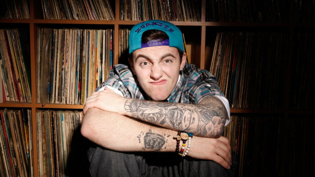 Mac Miller's Relaxed Stance amidst a Celestial Bookshelf: A QHD Wallpaper Masterpiece