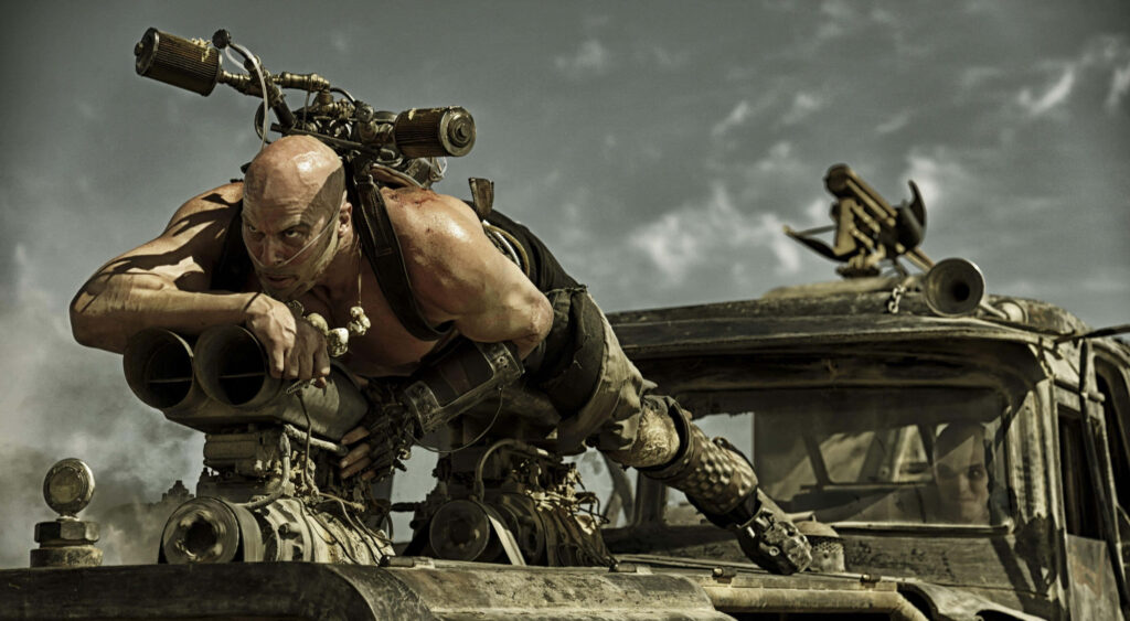 Mad Max Fury Road action scene with fierce character on armored vehicle in chaotic desert dust Wallpaper