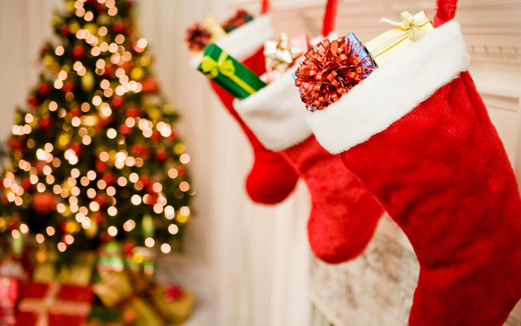 Festive Fireplace Delights: Three Adorned Stockings Overflowing with Gifts beside a Breathtakingly Decorated Christmas Tree Wallpaper