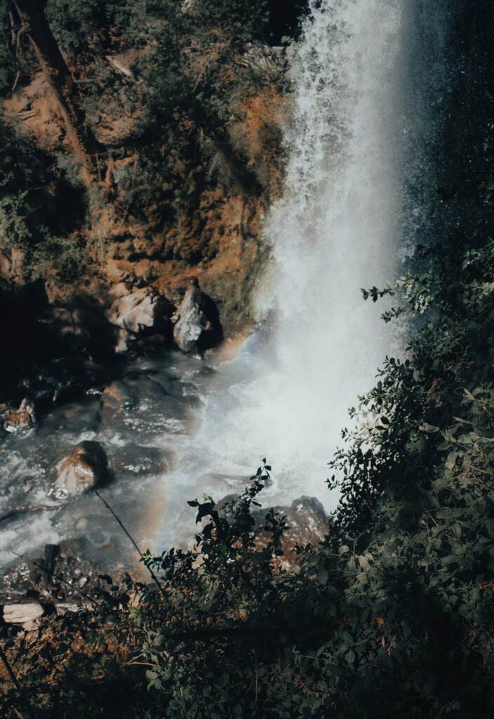 Waterfall cascading down cliff with mist and rainbow over smooth stones surrounded by green foliage Wallpaper